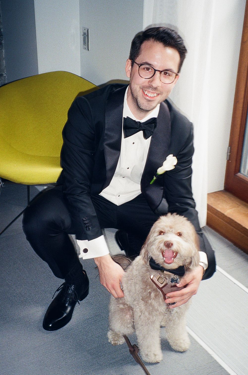 Man in tuxedo with bow tie crouching beside fluffy dog wearing bow tie collar, smiling indoors.