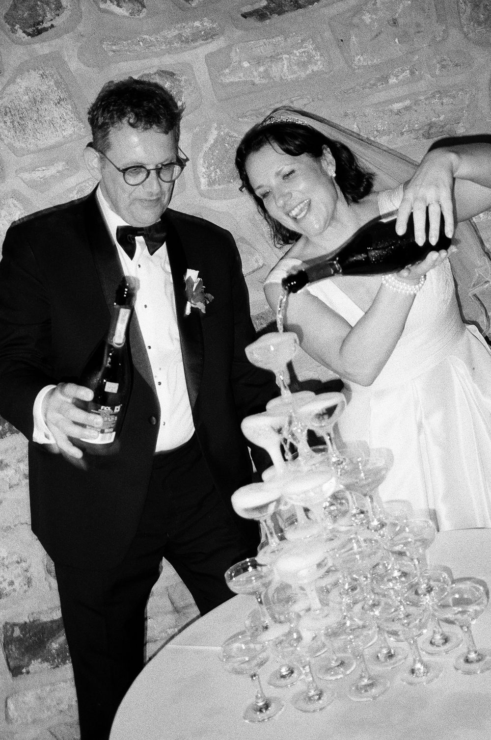 Black and white photo of a bride and groom pouring champagne tower at their wedding reception.