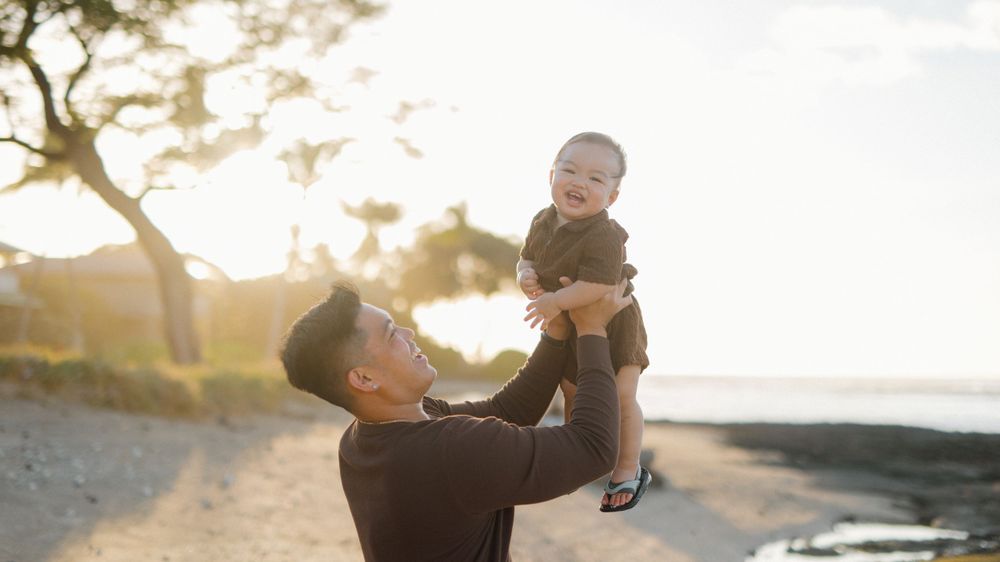 Father lifting his 1-year-old son in the air on a white sand beach at Puako, Hawaii, sharing a joyful moment.