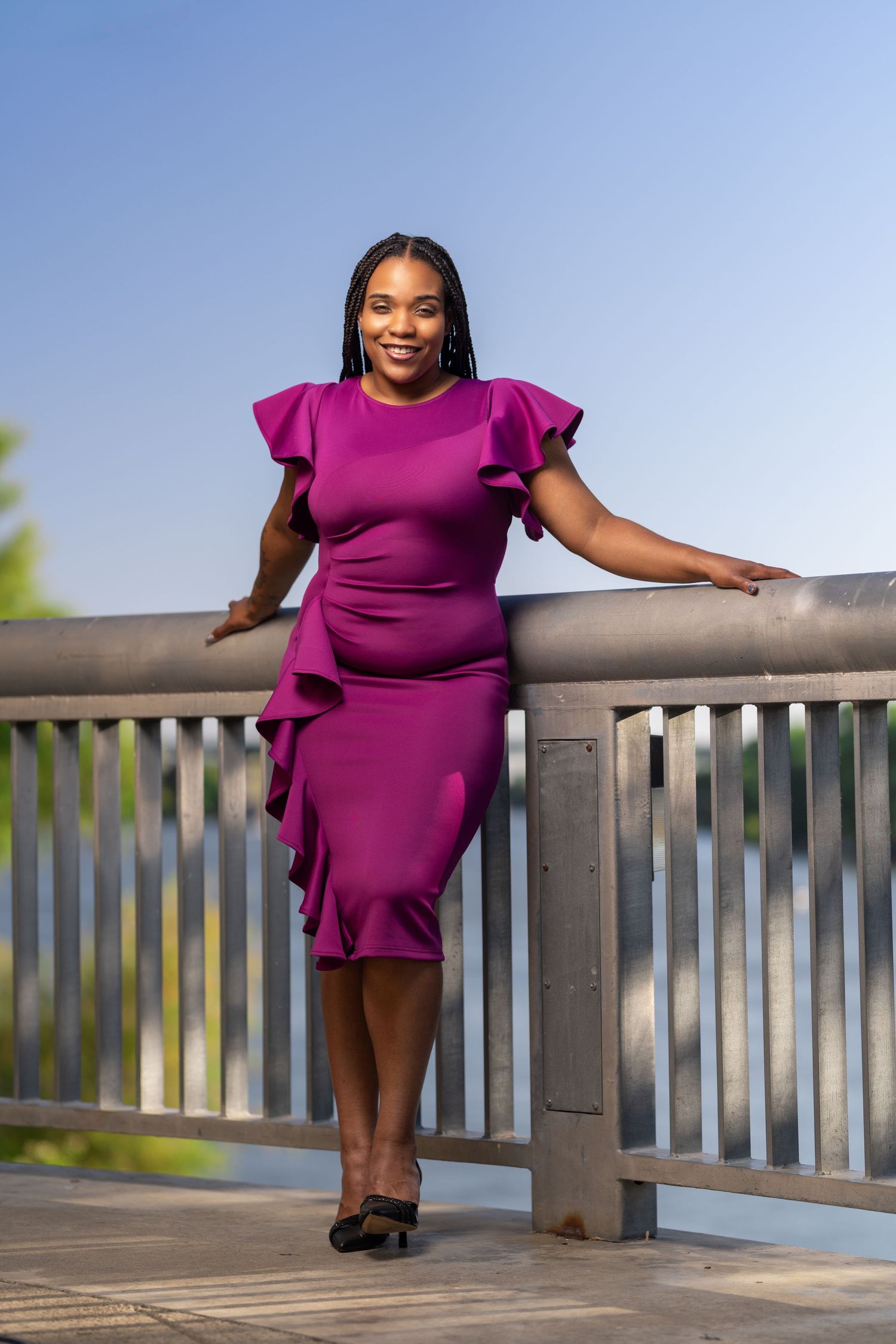 A person in a vibrant purple dress poses with confidence on a wooden deck railing against a bright sky backdrop.
