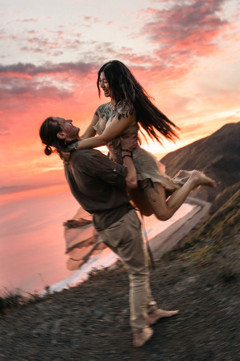 Couple dancing joyfully at sunset near coastal cliffs, woman lifted in air, vibrant pink and orange sky backdrop.