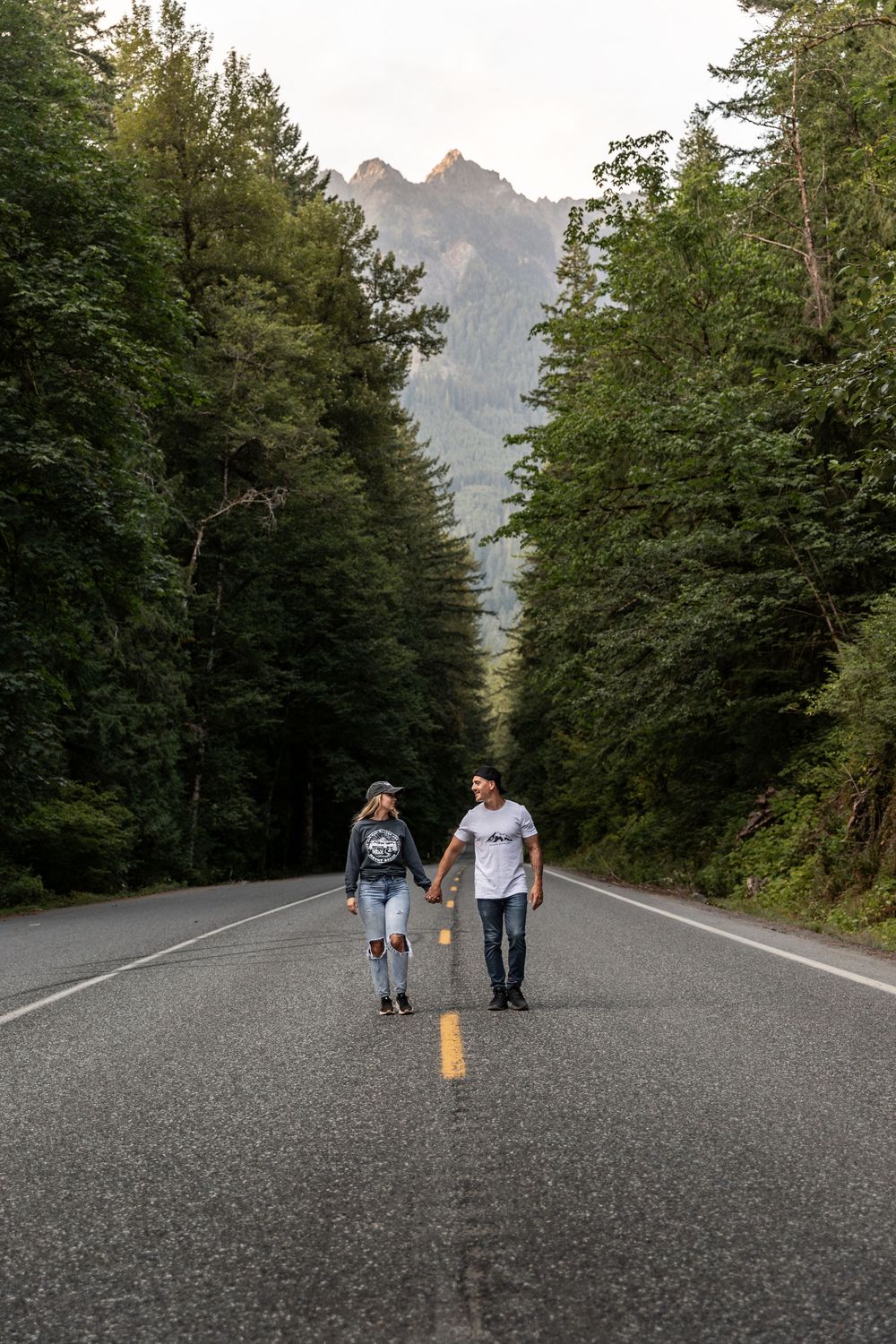 Couple walking down an empty mountain road surrounded by tall evergreen trees and distant mountain peaks.