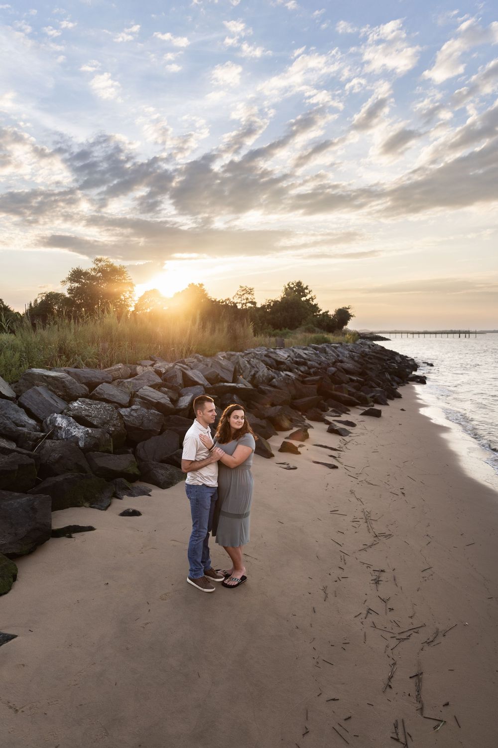 Couple embraces on sunset beach with rocky jetty and dramatic cloudy sky in background. Woodland Beach Engagement Photos