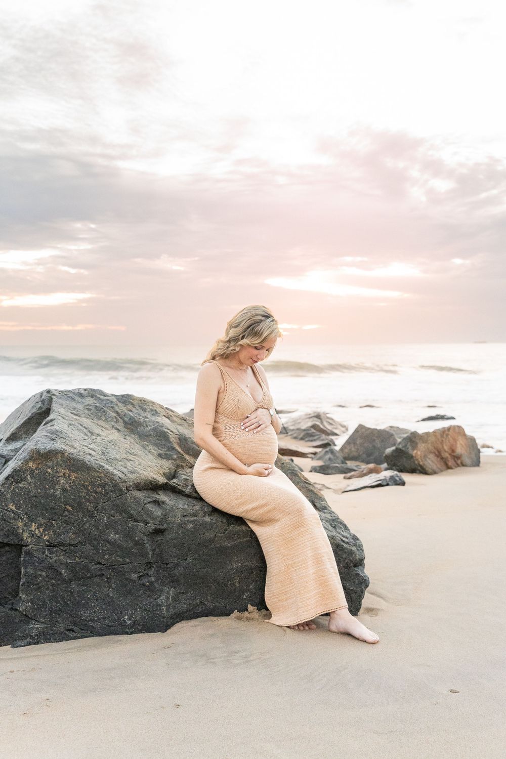 A person in a peachy dress sits gracefully on dark coastal rocks during a dreamy beach sunset.