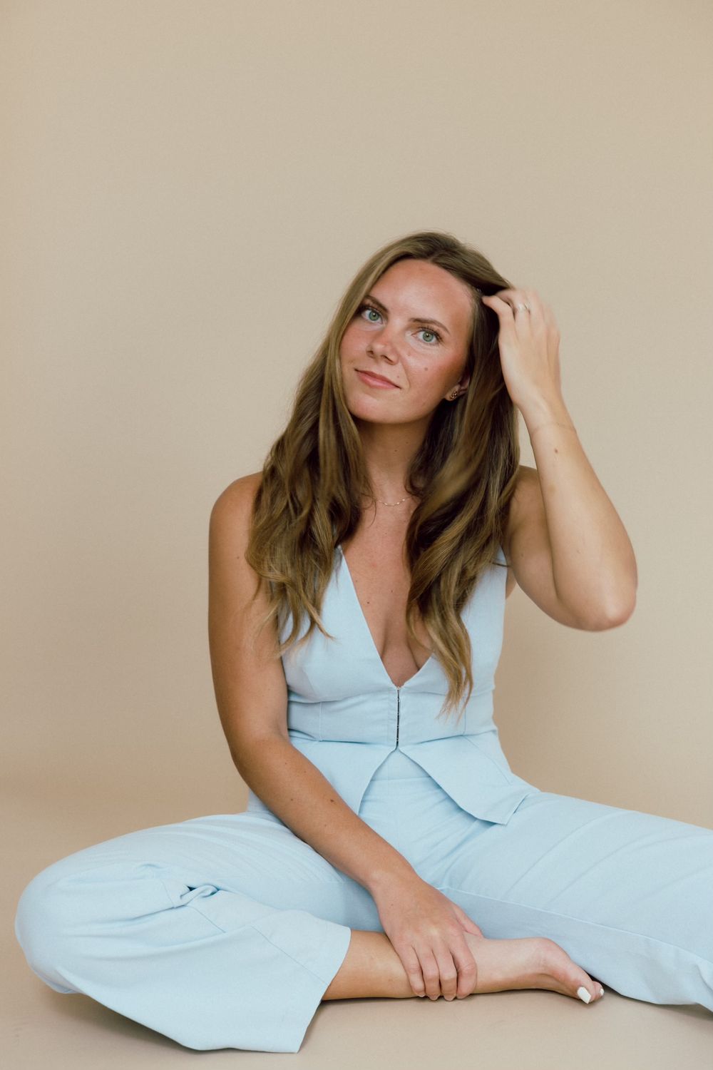 Woman in light blue jumpsuit sitting casually against neutral background.