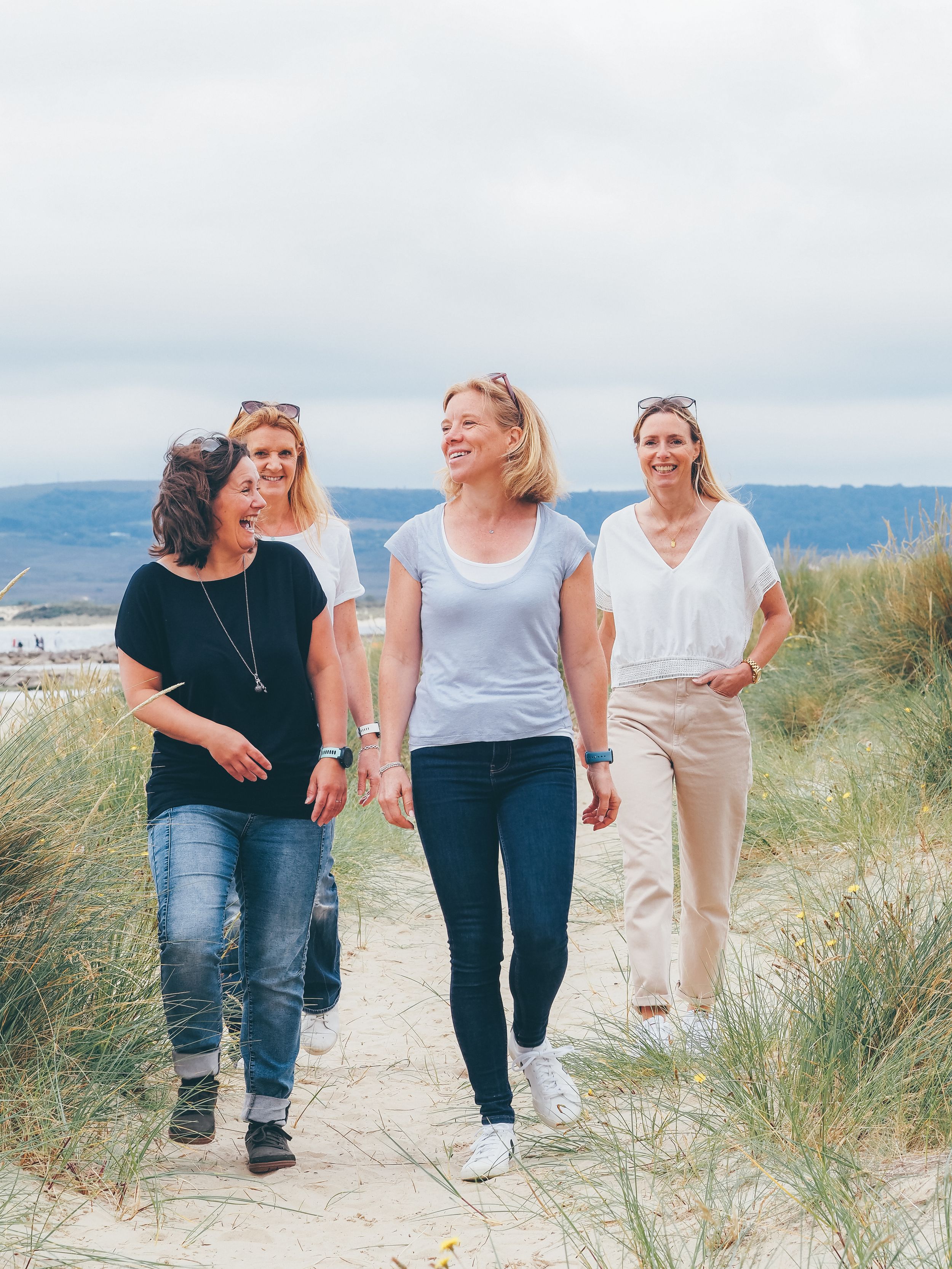 Three friends walking and laughing together along a sandy beach path with coastal mountains in the background.