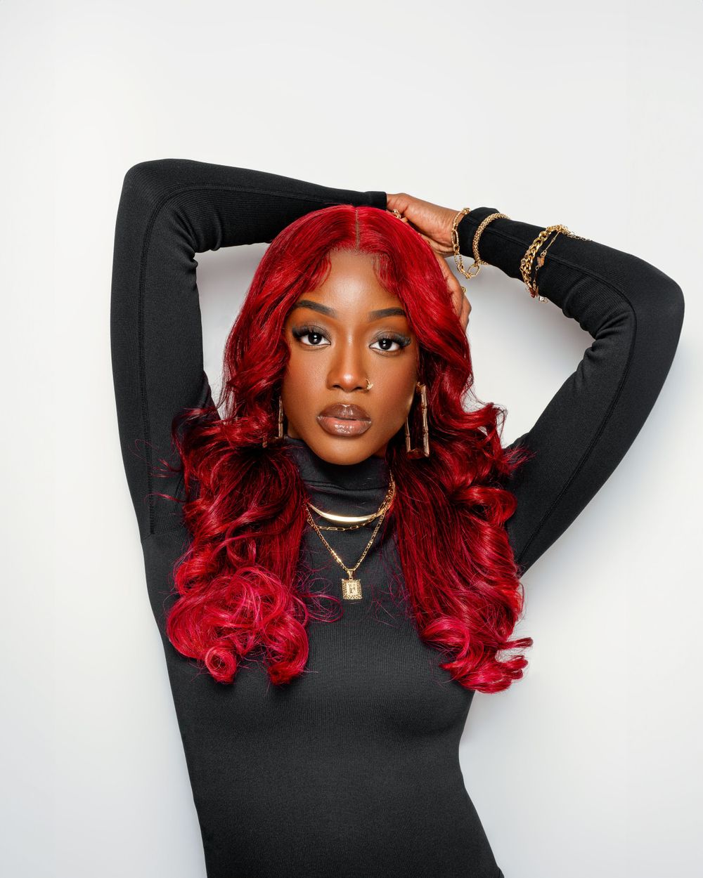 Portrait of woman with vibrant red curls wearing black turtleneck and layered gold necklaces.