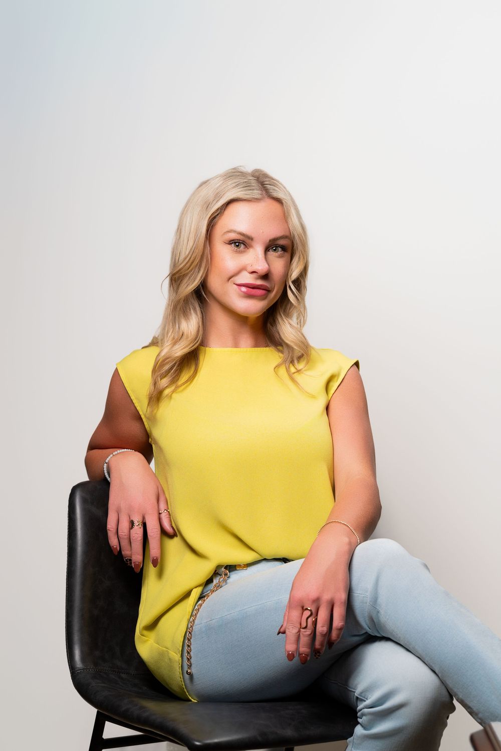 A person in a yellow sleeveless top and light jeans sits casually on a black chair against a white background.