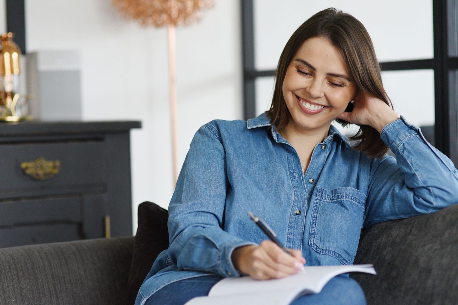 Someone wearing a denim shirt sits comfortably on a couch while writing in a notebook and smiling. Shots taken on a brand photoshoot.