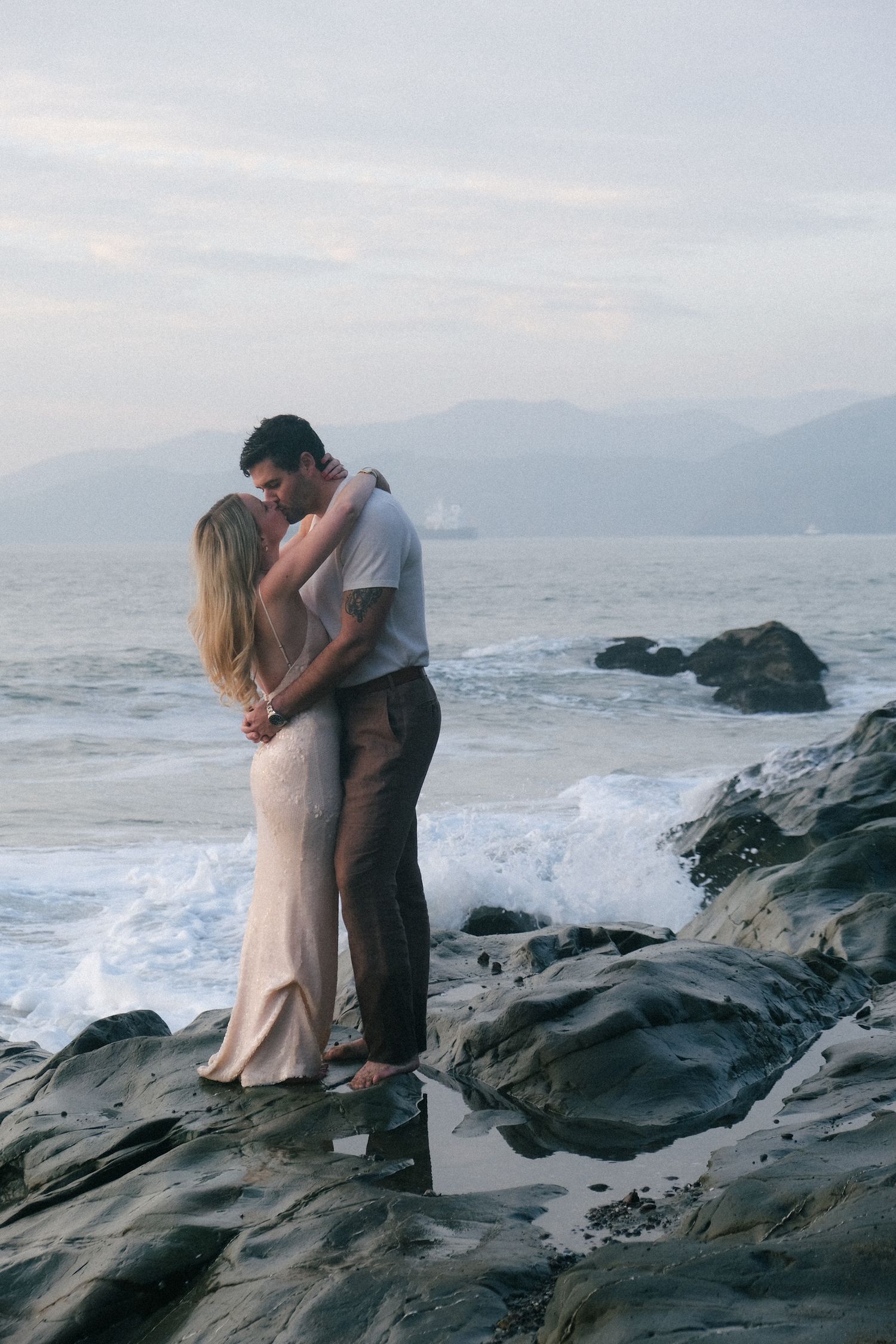 A romantic couple embraces on rocky coastal cliffs by the ocean.