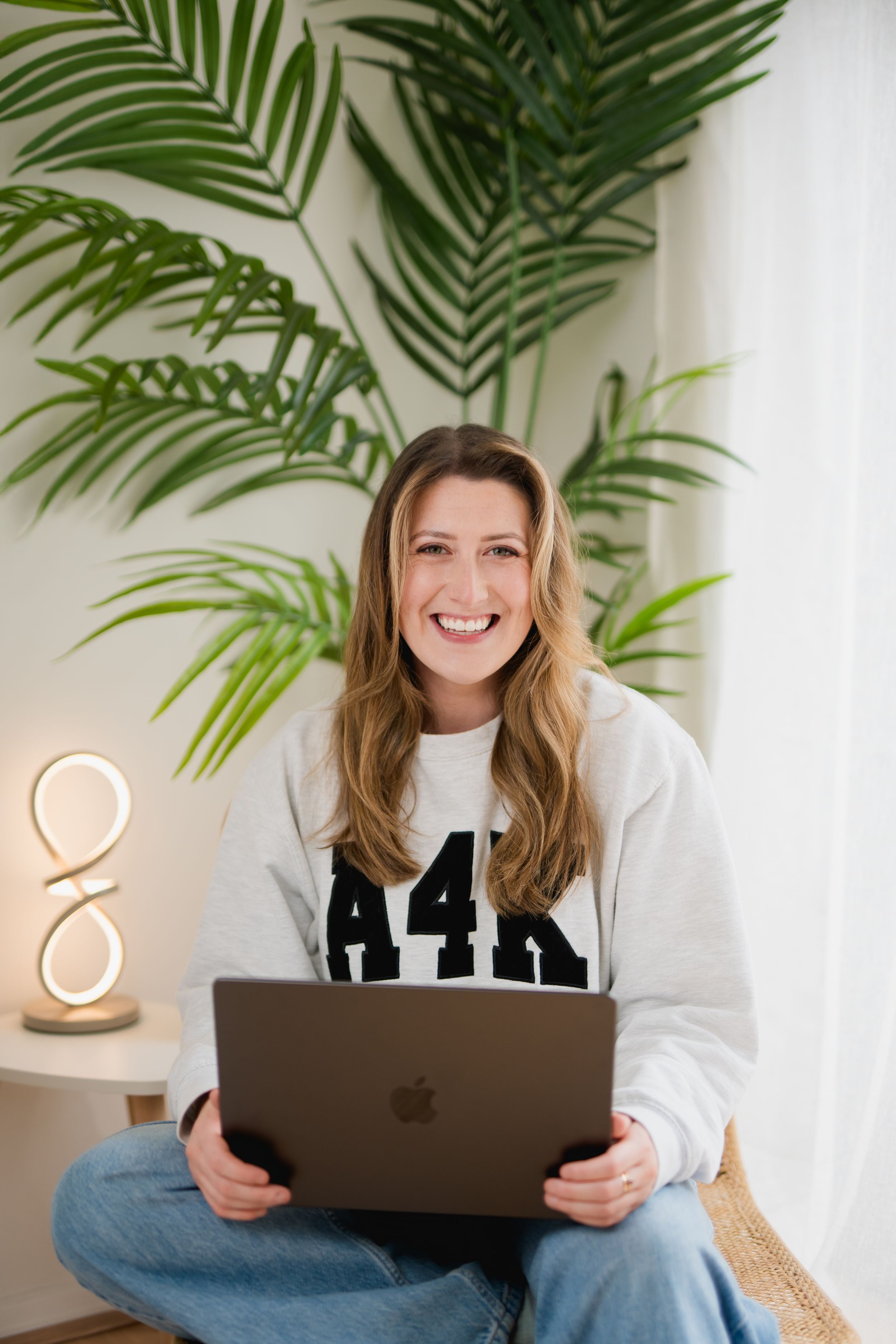 Remote worker in cozy home office with laptop and palm plants sits comfortably on white couch.