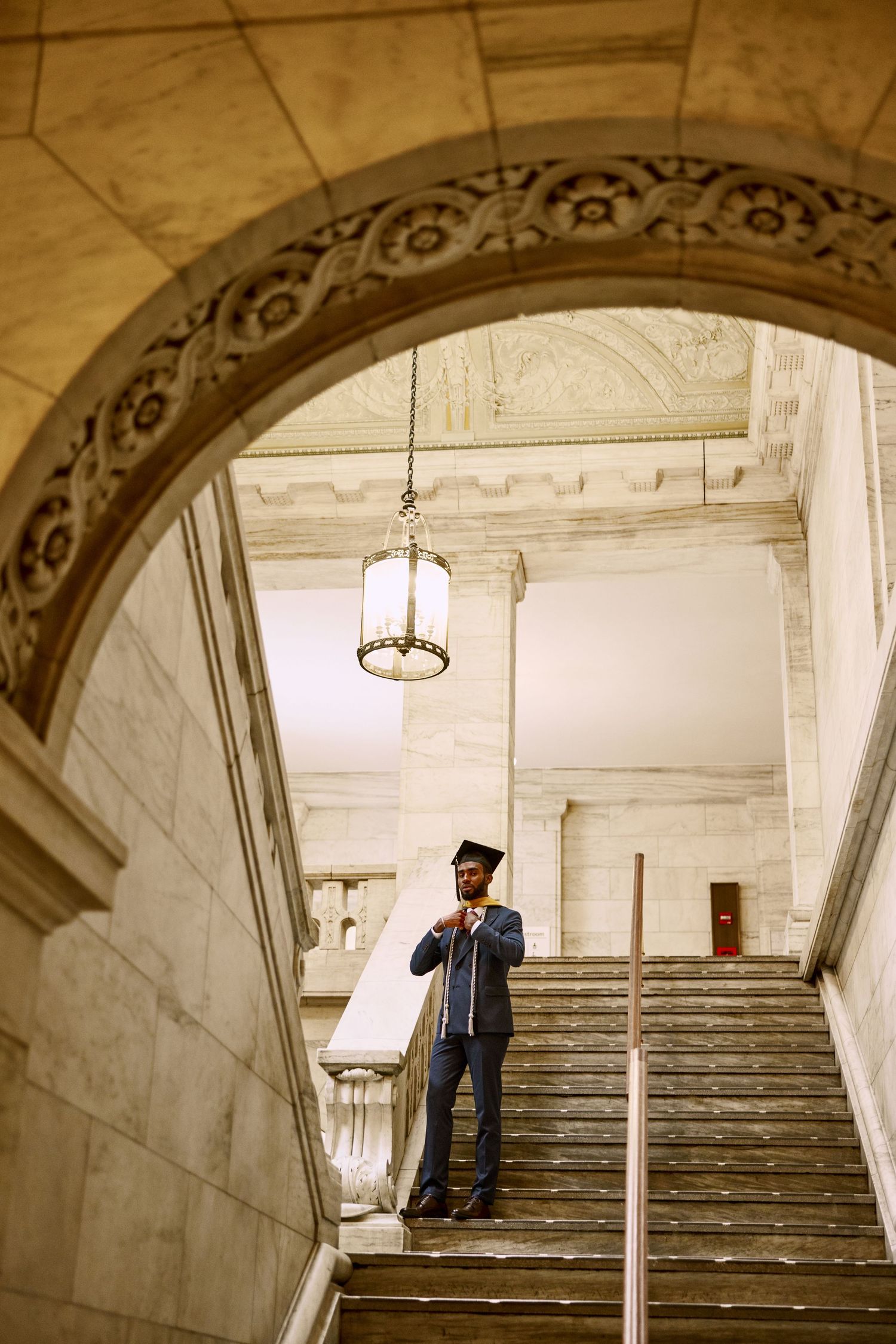Graduate student in cap and gown stands on grand marble staircase under ornate archway in historic building.