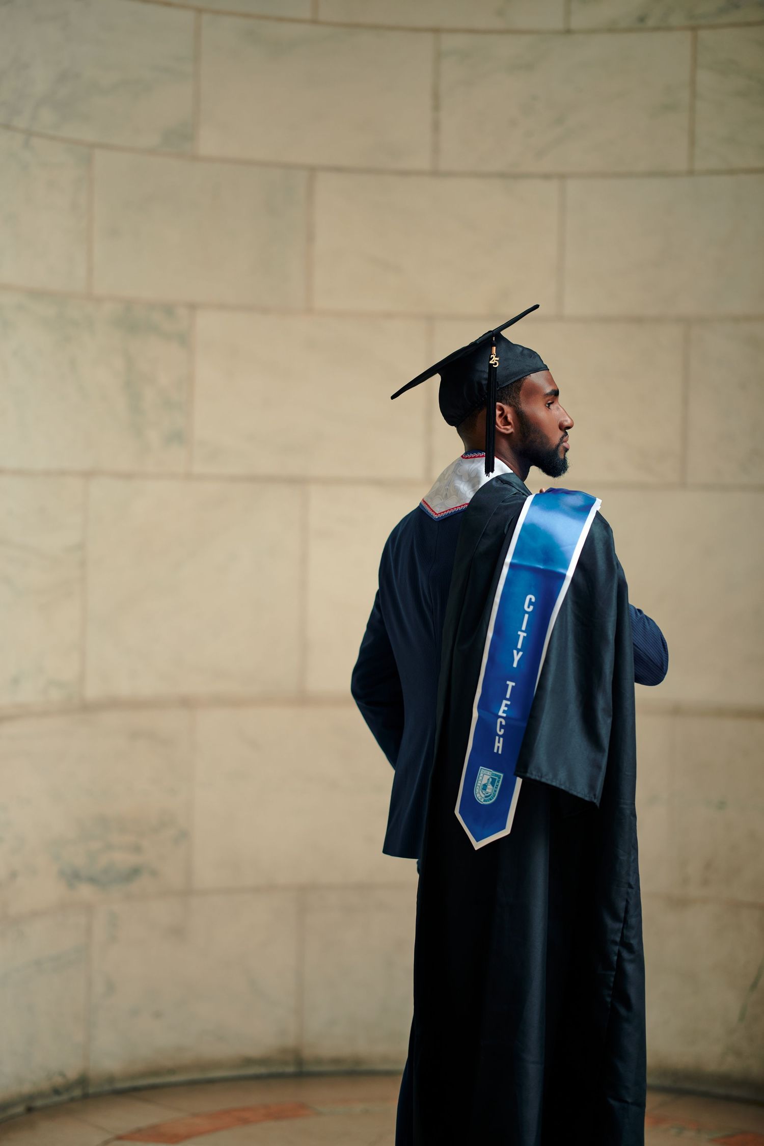 A graduate in black cap and gown with a blue and white graduation sash stands against a light stone wall.
