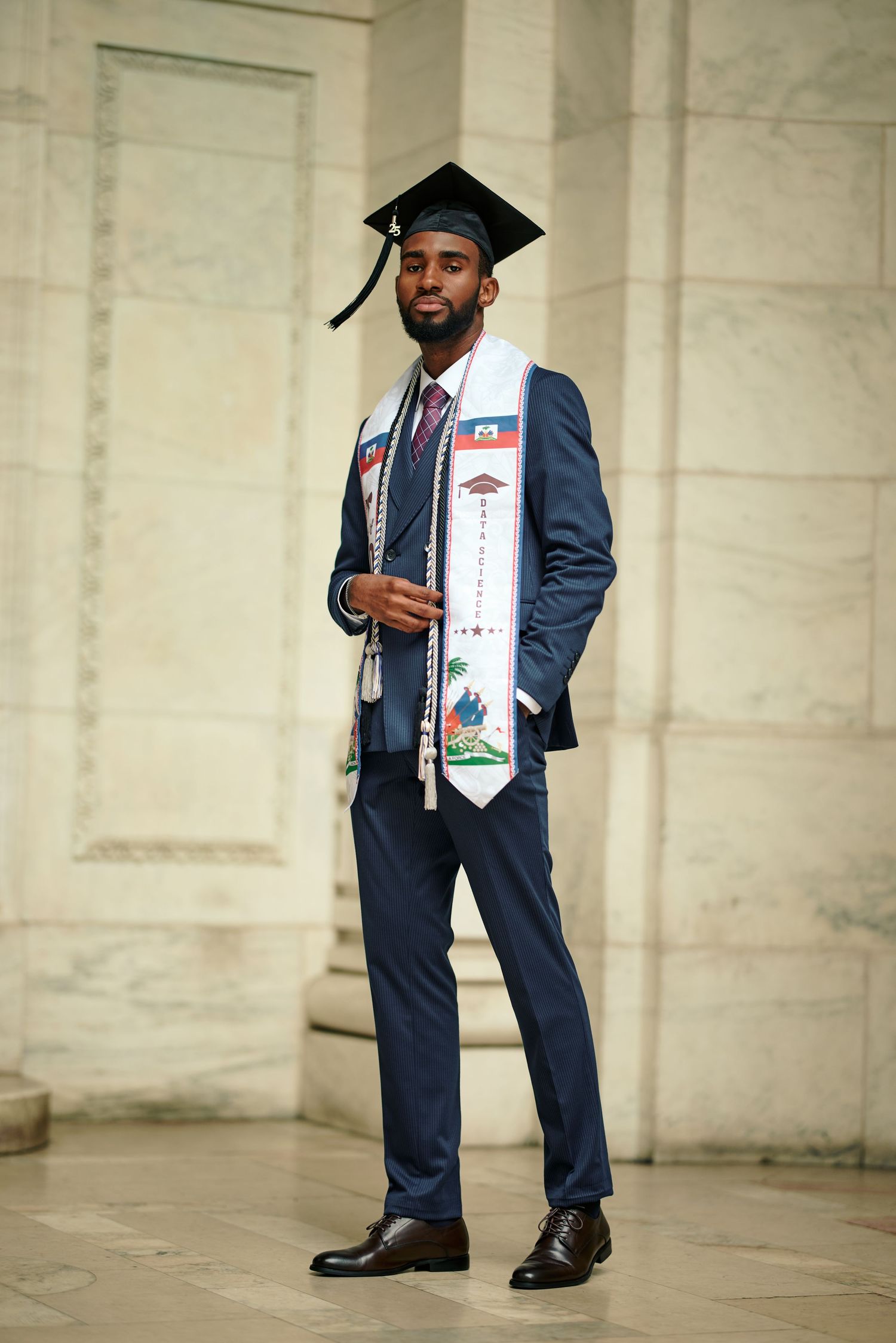 Graduate in navy blue suit and graduation cap poses confidently with decorated stole and diploma during commencement ceremony.