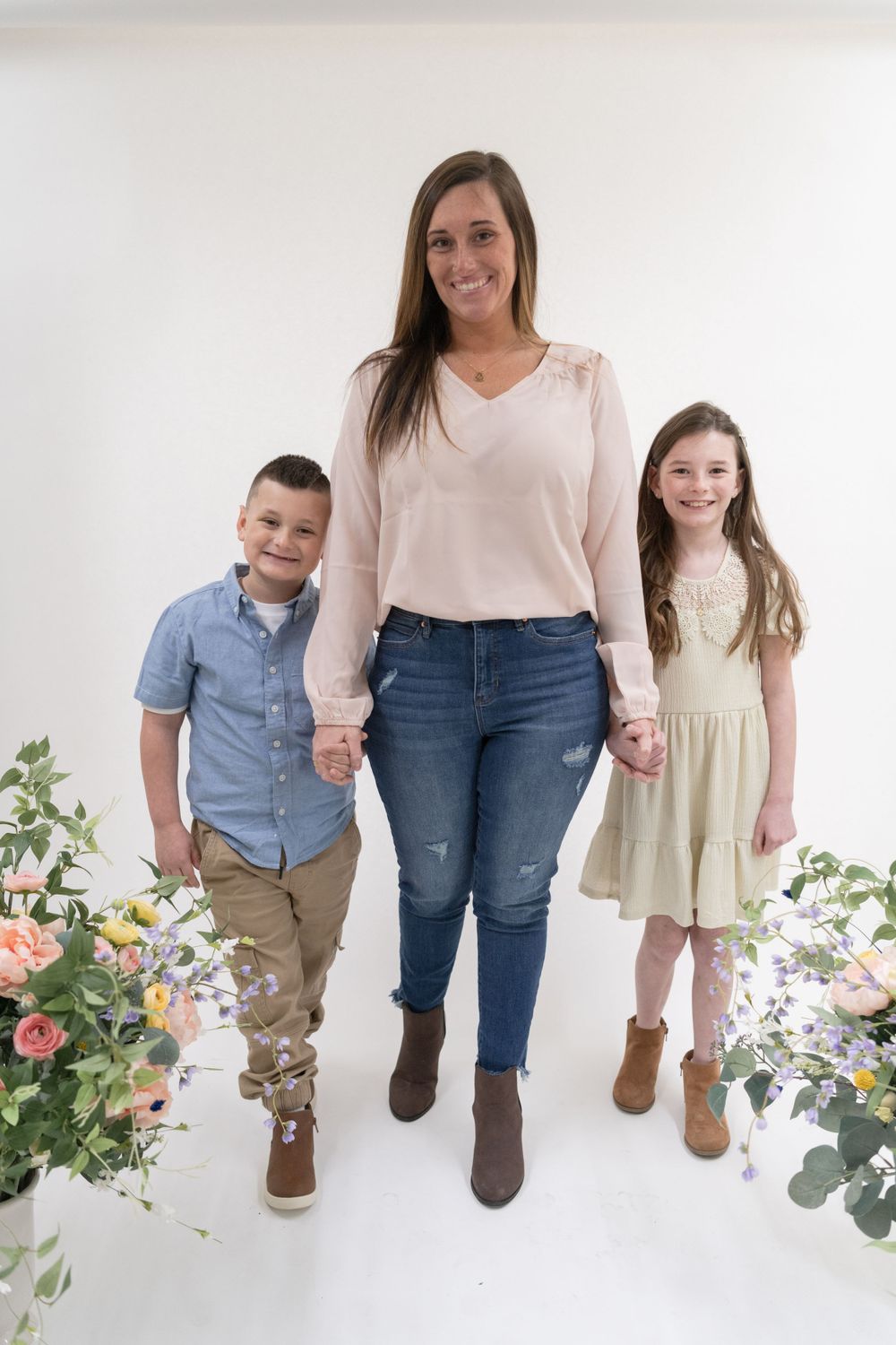 Family portrait with mother and two children standing between flower arrangements on white background.
