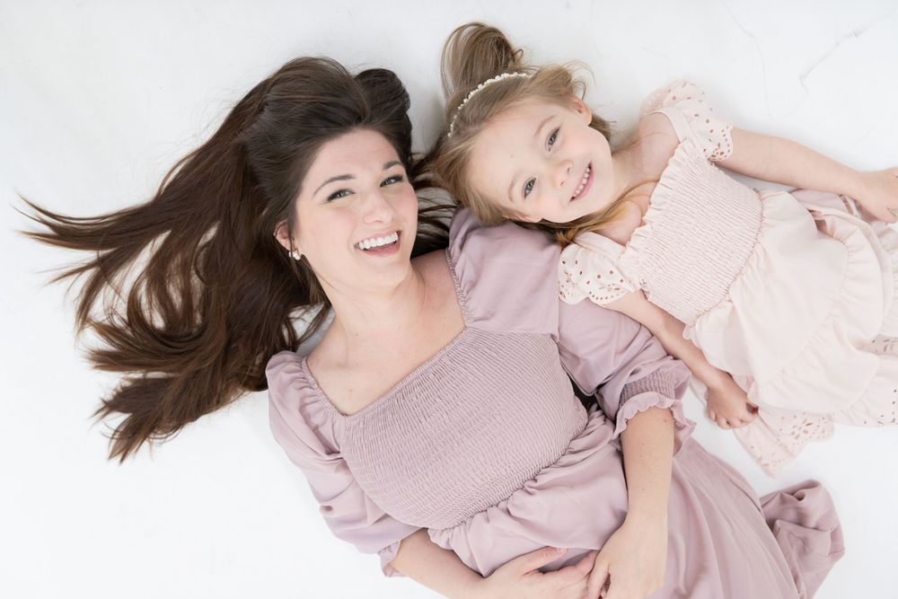 Parent and child lying on white background wearing pink dresses while smiling and embracing.
