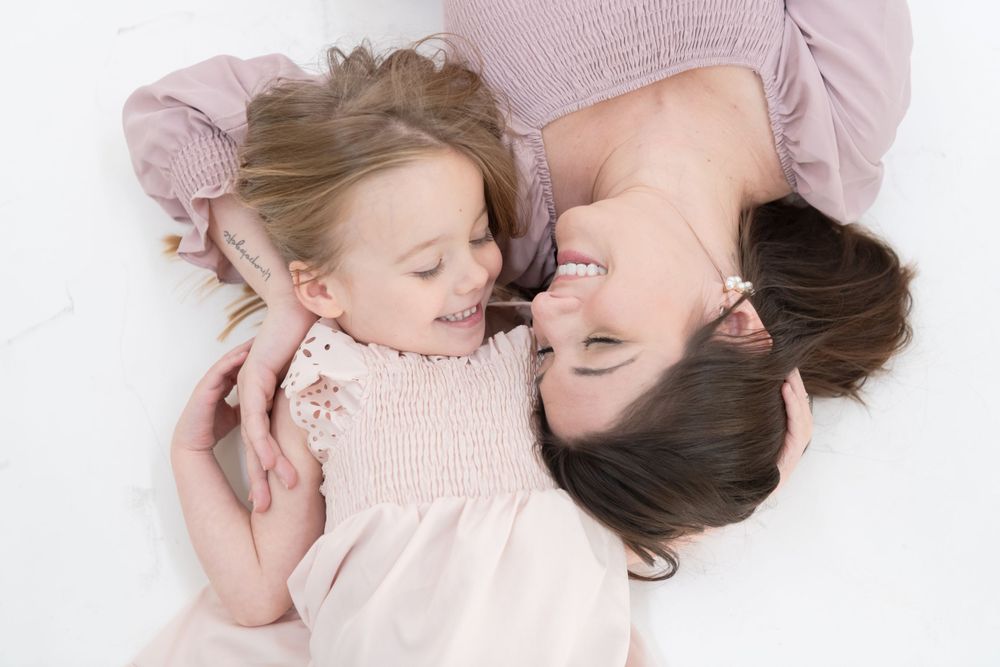 Parent and child in matching soft pink dresses lying close together on white backdrop.