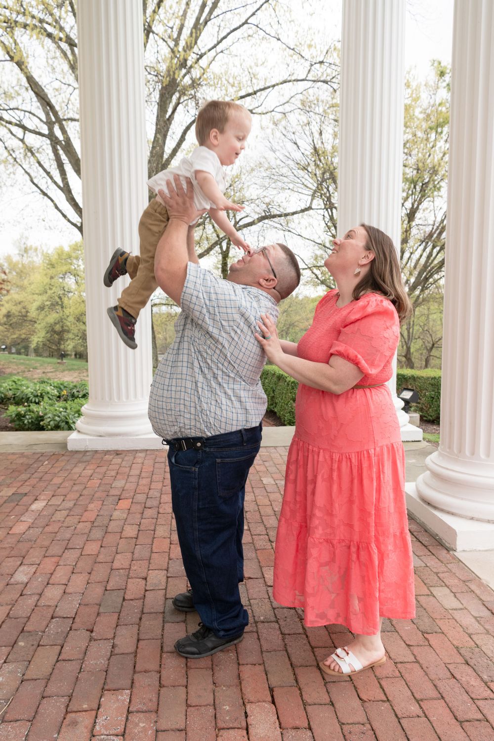 Joyful outdoor family moment with parents lifting young child by brick columns.