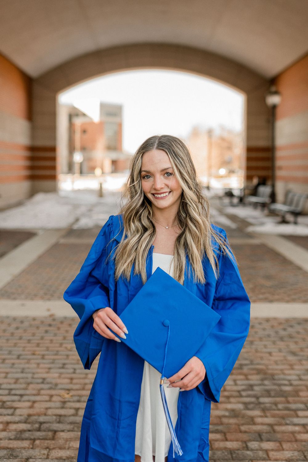 Smiling graduate in blue cap and gown holding diploma outdoors near brick archway.
