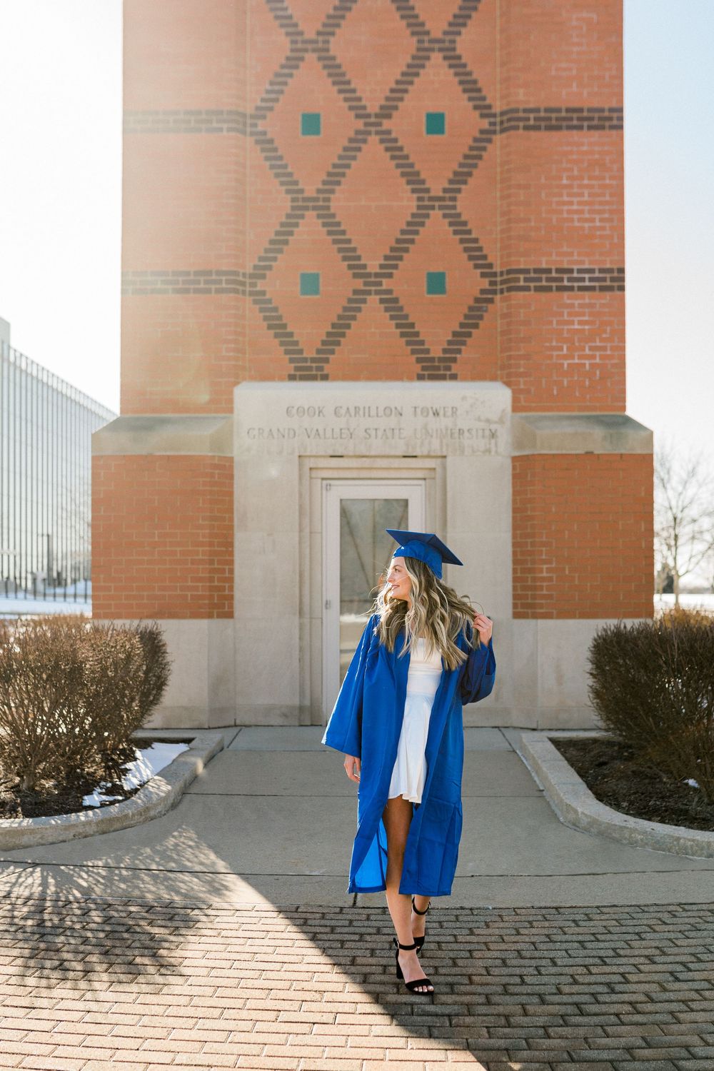 A woman in a blue graduation cap and gown poses in front of Cook Carillon Tower at Grand Valley State University.