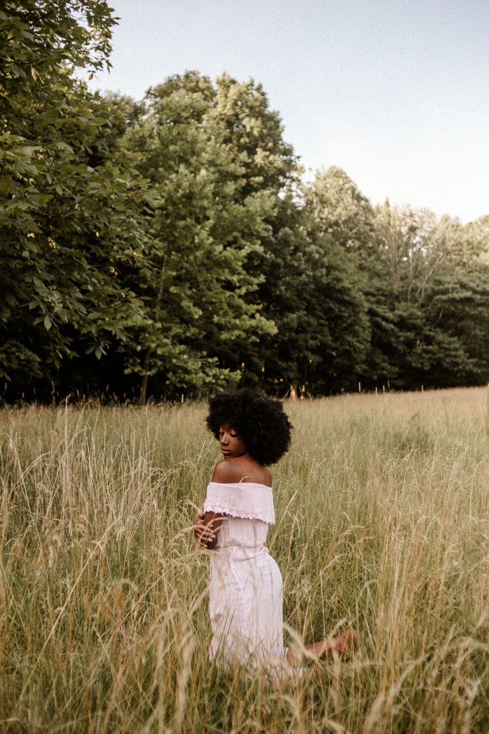 Woman in white off-shoulder dress standing in tall grass field with lush green trees in background.