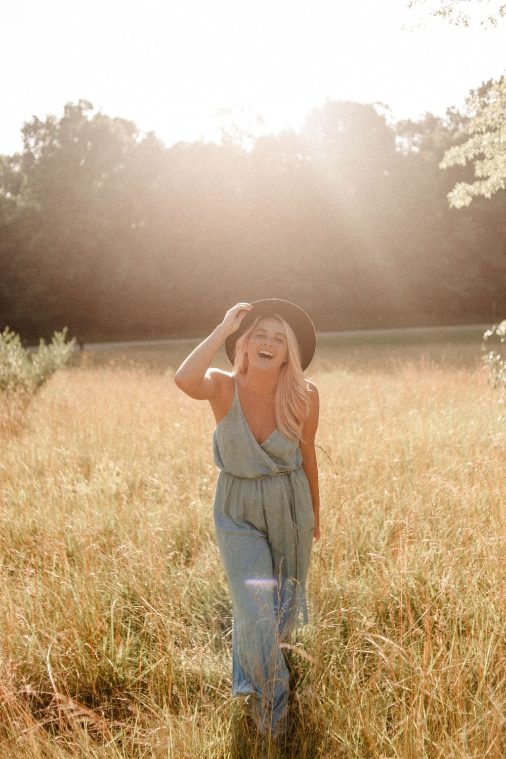Smiling blonde woman in green jumpsuit and black hat standing in a sunlit golden field.