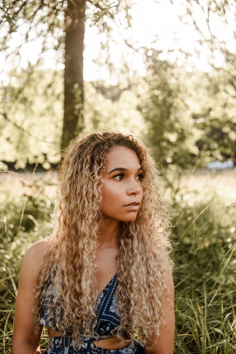 Young woman with long curly blonde hair sitting outdoors among lush green trees and grass.