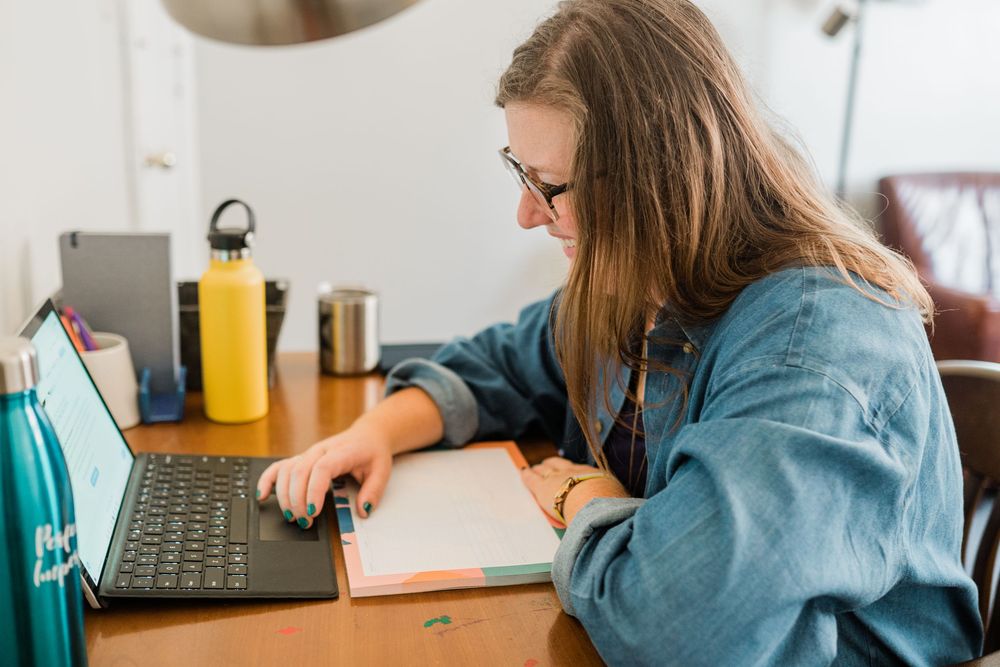 Young woman with glasses studying at desk, using laptop and taking notes in notebook at home.