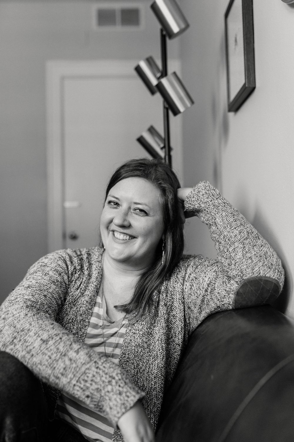 Black and white portrait of a smiling woman relaxing on a sofa in a cozy living room with a modern floor lamp.