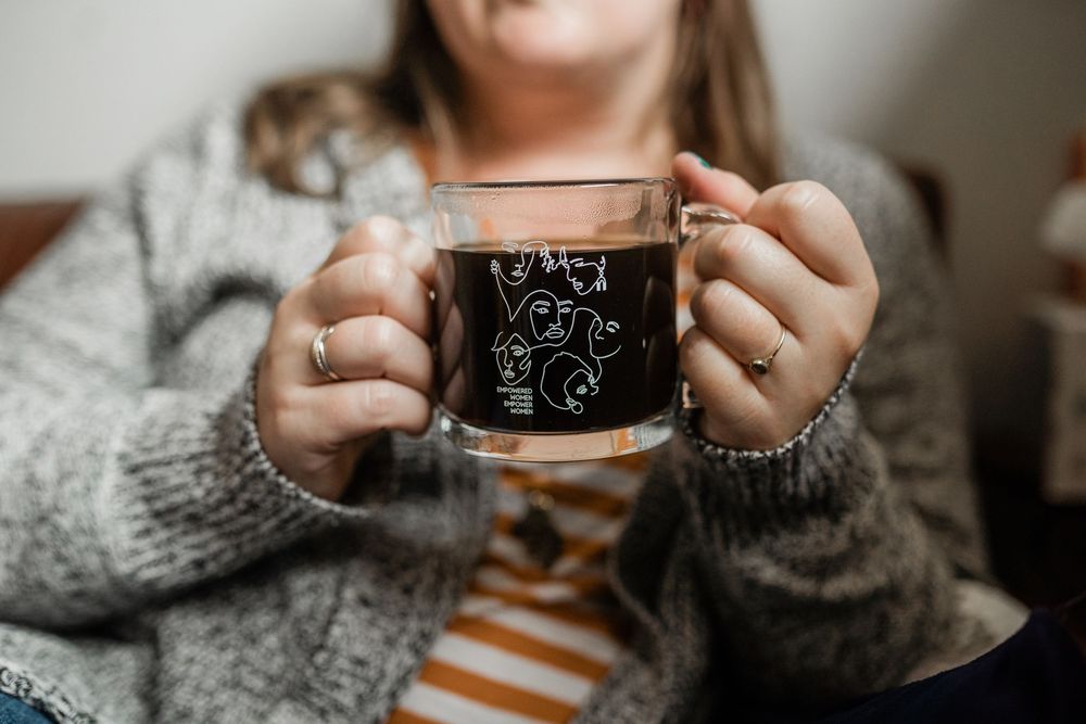 Woman in gray knit sweater holding a clear glass mug with black ghost design containing dark tea or coffee.