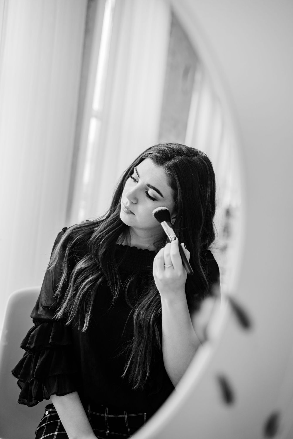 Black and white photo of a young woman applying makeup with a brush in front of a round mirror.