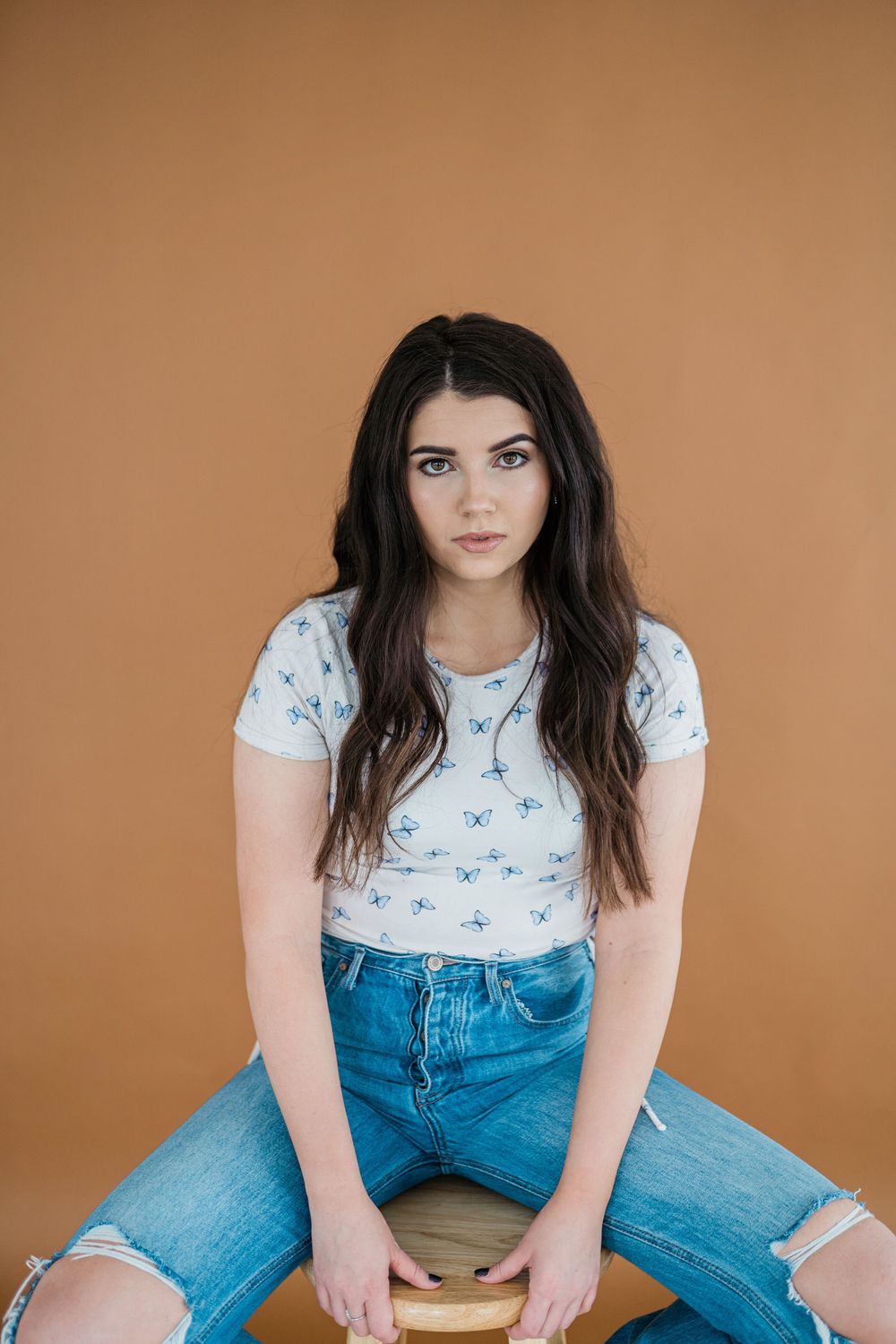 Young woman with long dark hair sits on stool wearing white patterned tee and ripped blue jeans against tan background.
