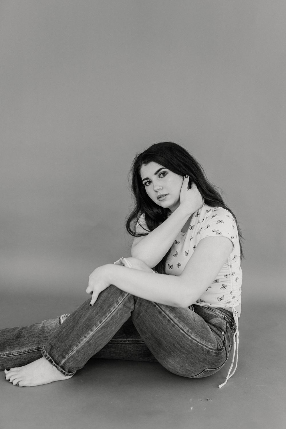 Black and white portrait of a young brunette woman sitting on the floor in jeans and a floral top, looking at the camera.