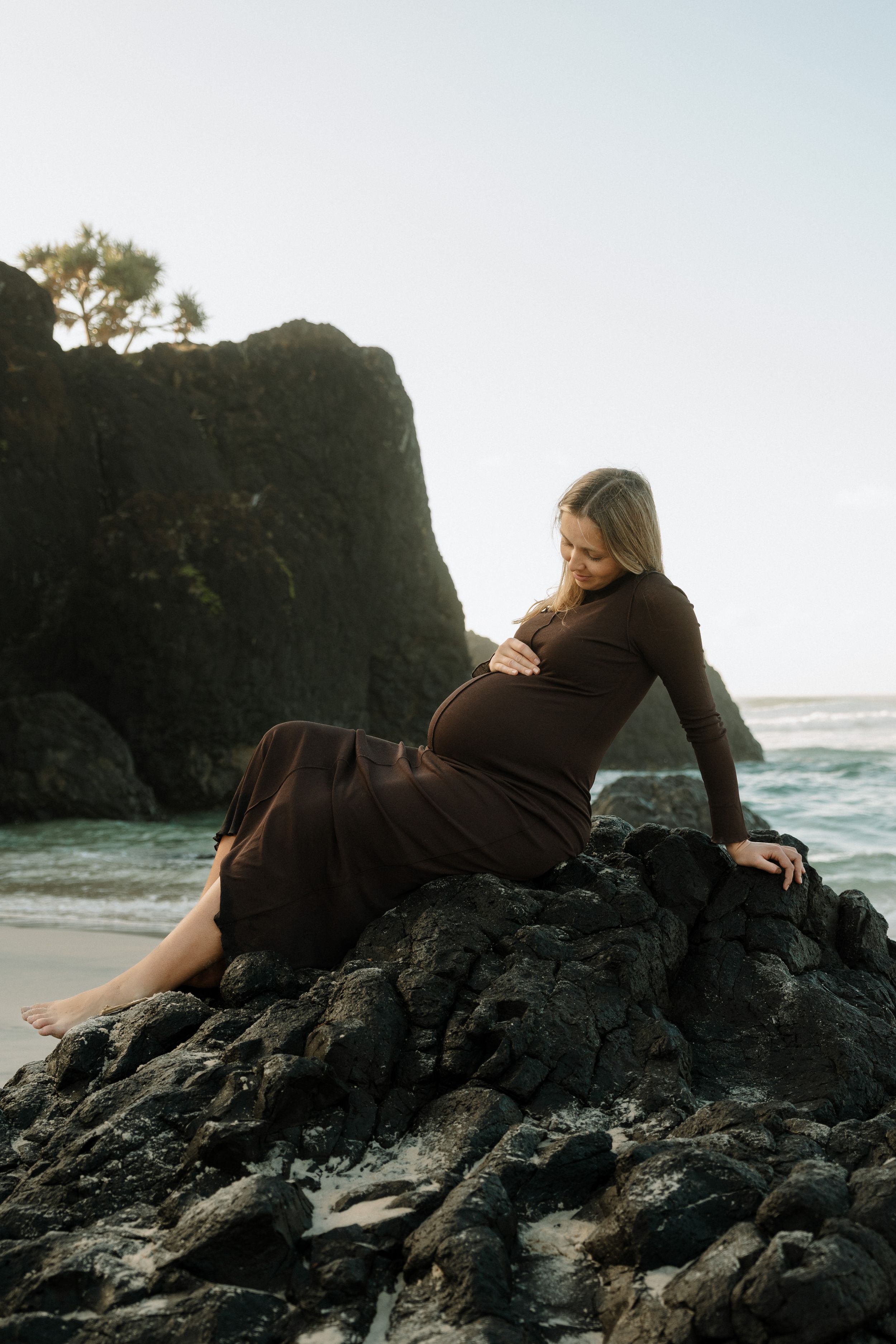 Maternity portrait on black volcanic rocks at sunset with ocean waves in background.