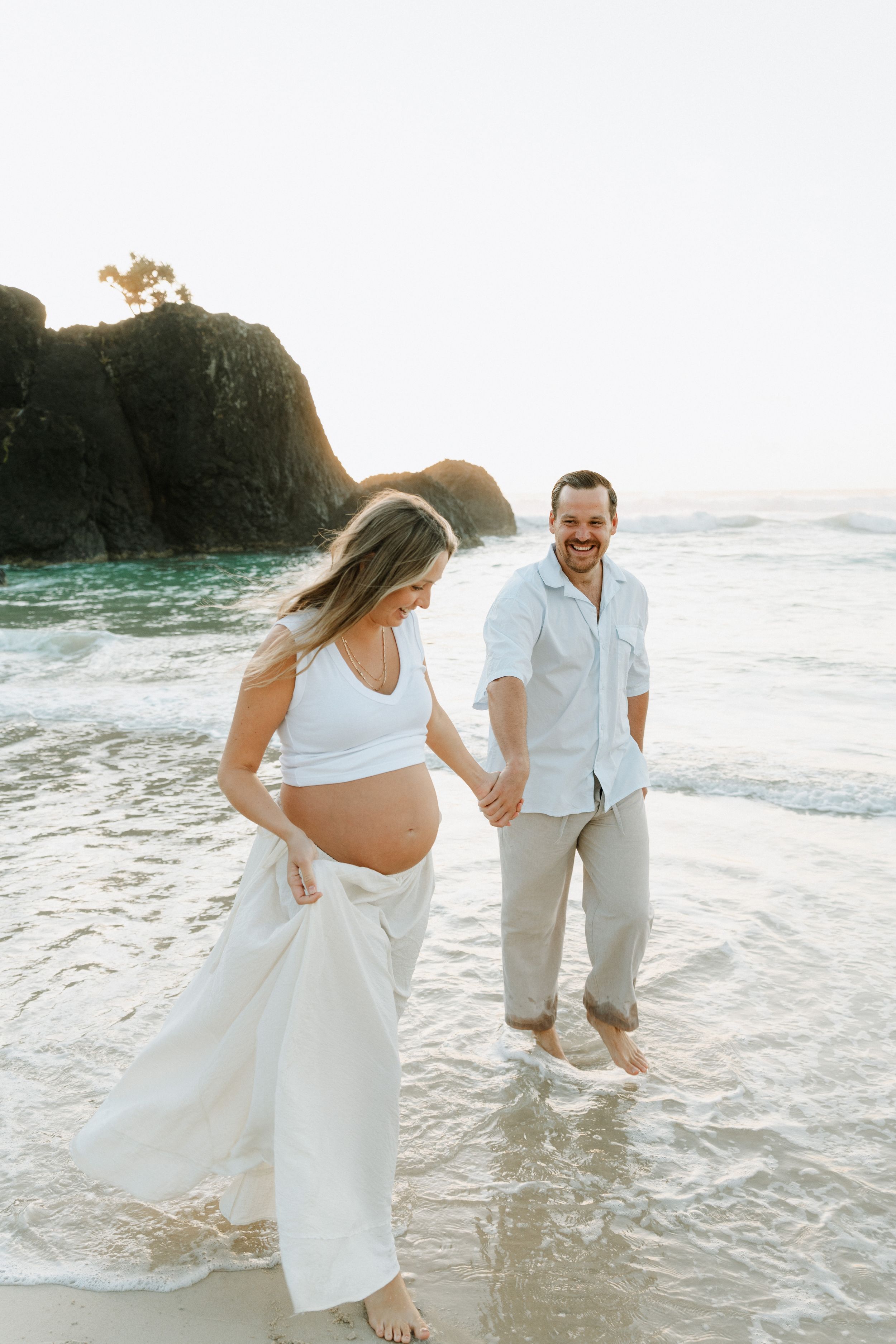 Couple playing in shallow ocean waves wearing all white outfits on sandy beach.