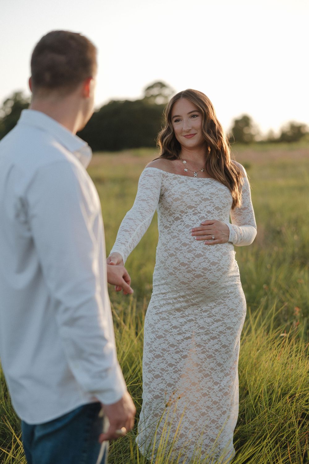 Pregnant woman in white lace dress holds hands with man in green field at golden hour sunset.
