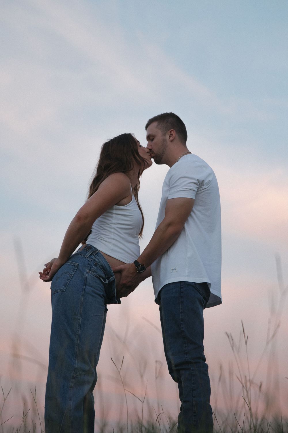Couple kissing romantically in a field at dusk, both wearing white tops and blue jeans under a pastel sky.