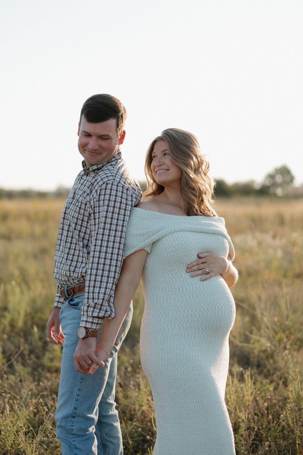 Couple shares intimate moment in field during sunset maternity session wearing plaid and white.