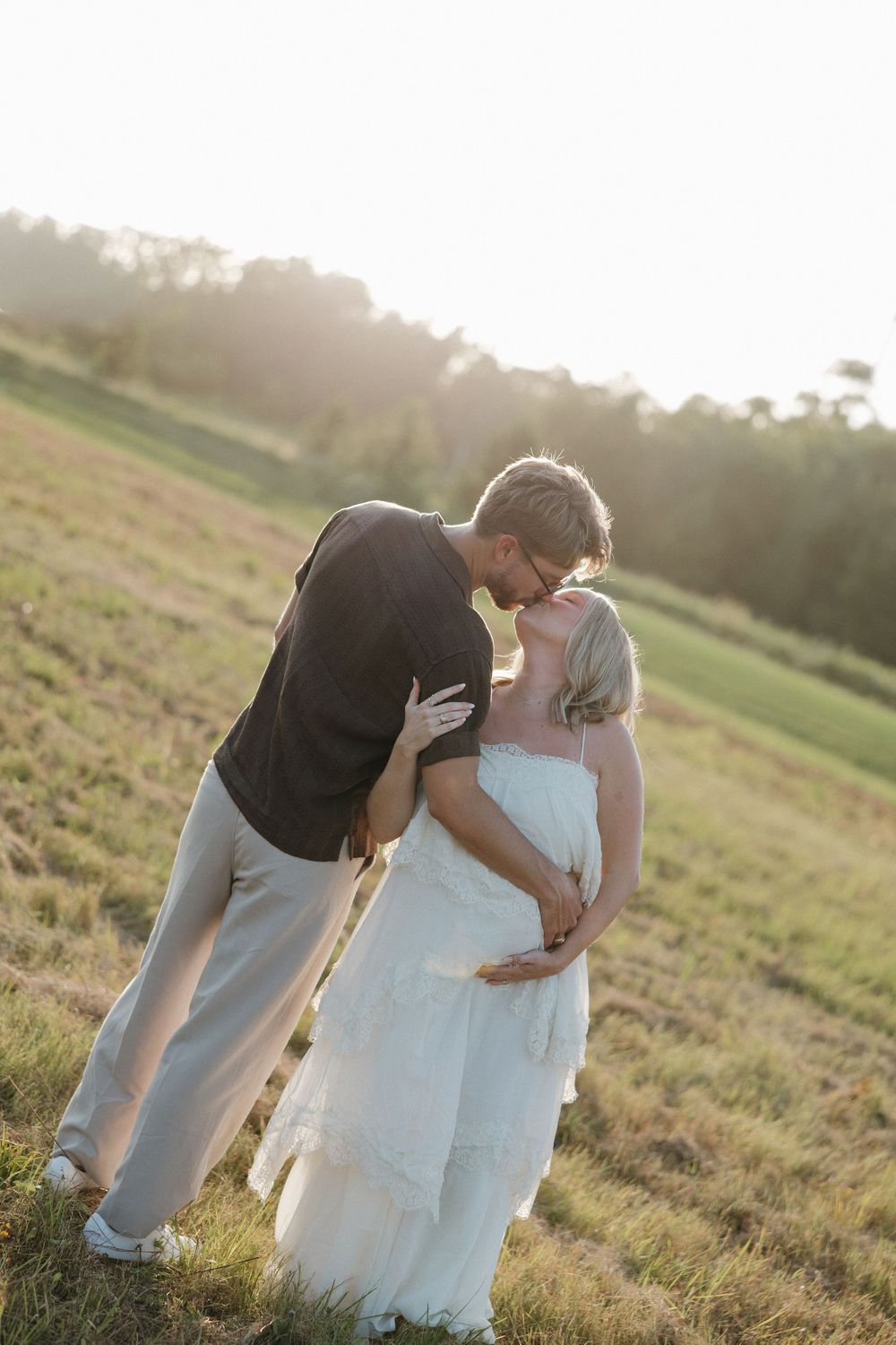 Couple sharing a tender kiss in a sunlit green field, woman in white dress with visible baby bump, golden hour lighting.