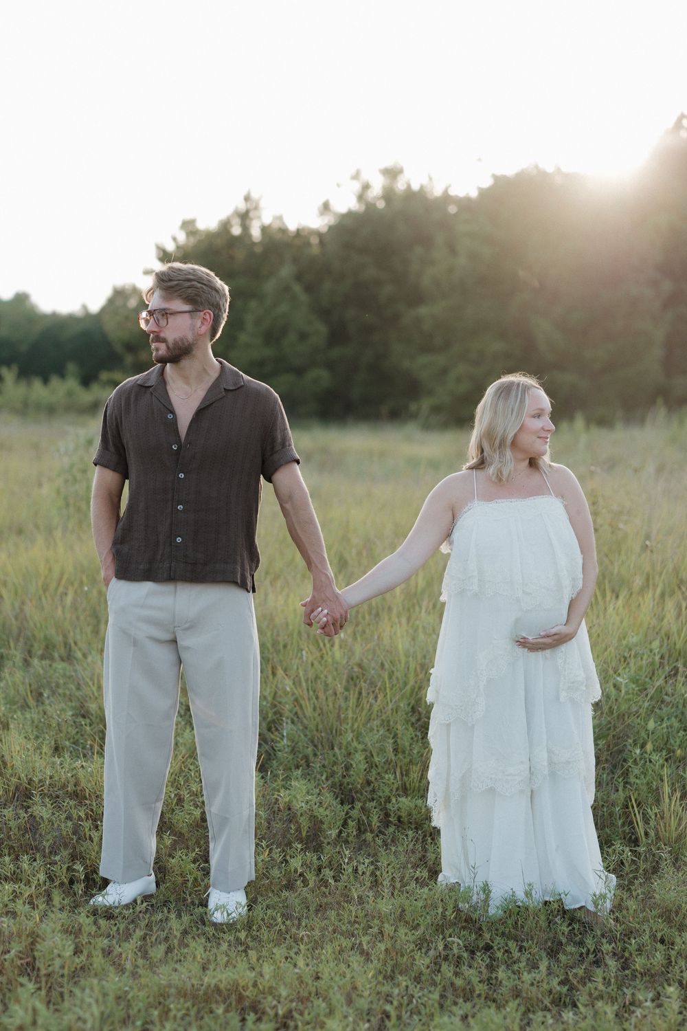 Pregnant couple holding hands in a sunlit field, woman in white dress touching belly, man in dark shirt and khakis.