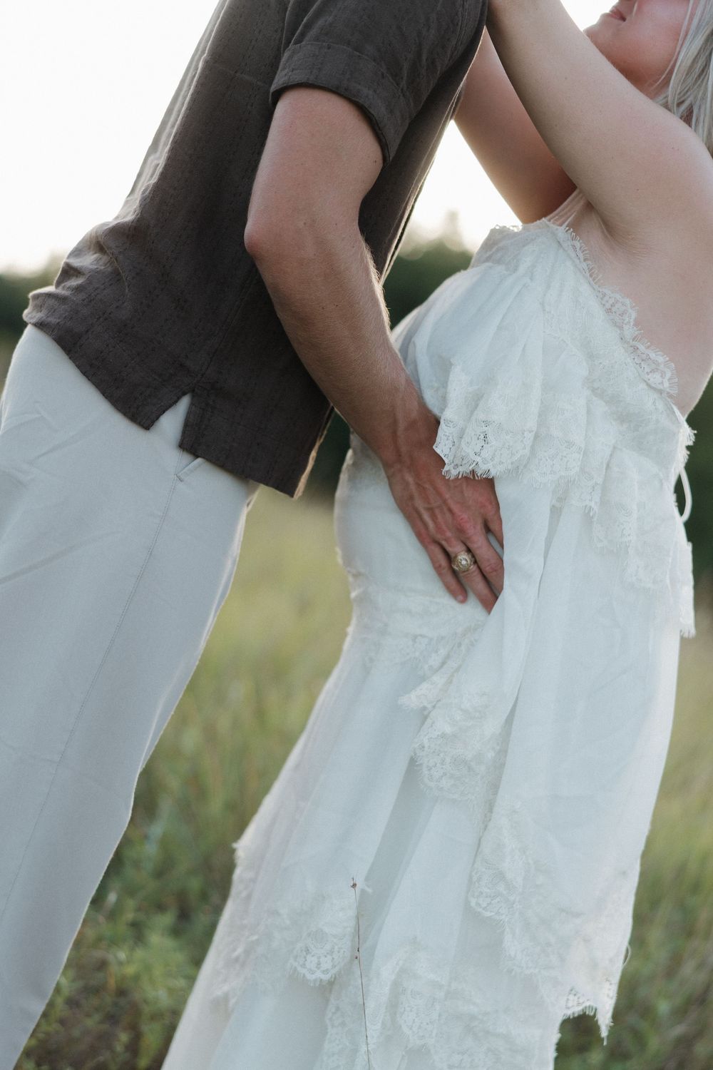 A couple in a field, man in gray shirt and white pants holding woman in white lace dress, wearing a ring.