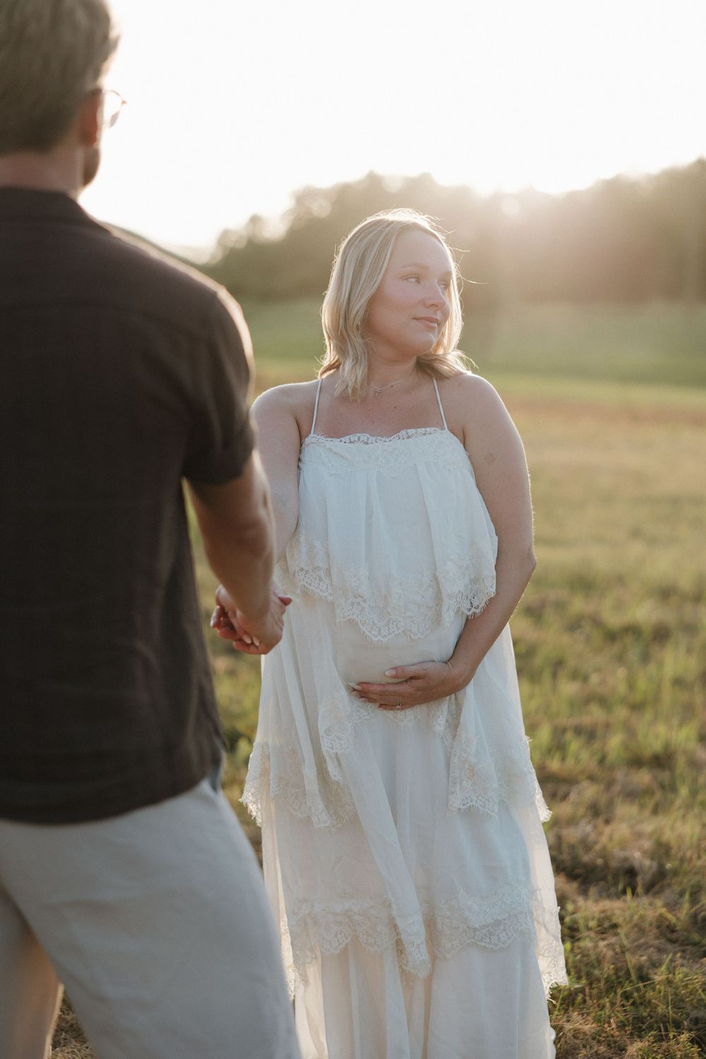 Pregnant woman in white lace dress holds belly at golden hour in a field, holding hands with a man.