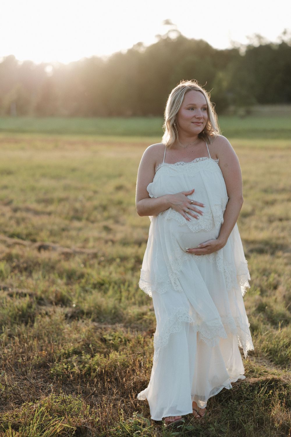 Pregnant woman in white dress holding belly in golden sunlit field at sunset.