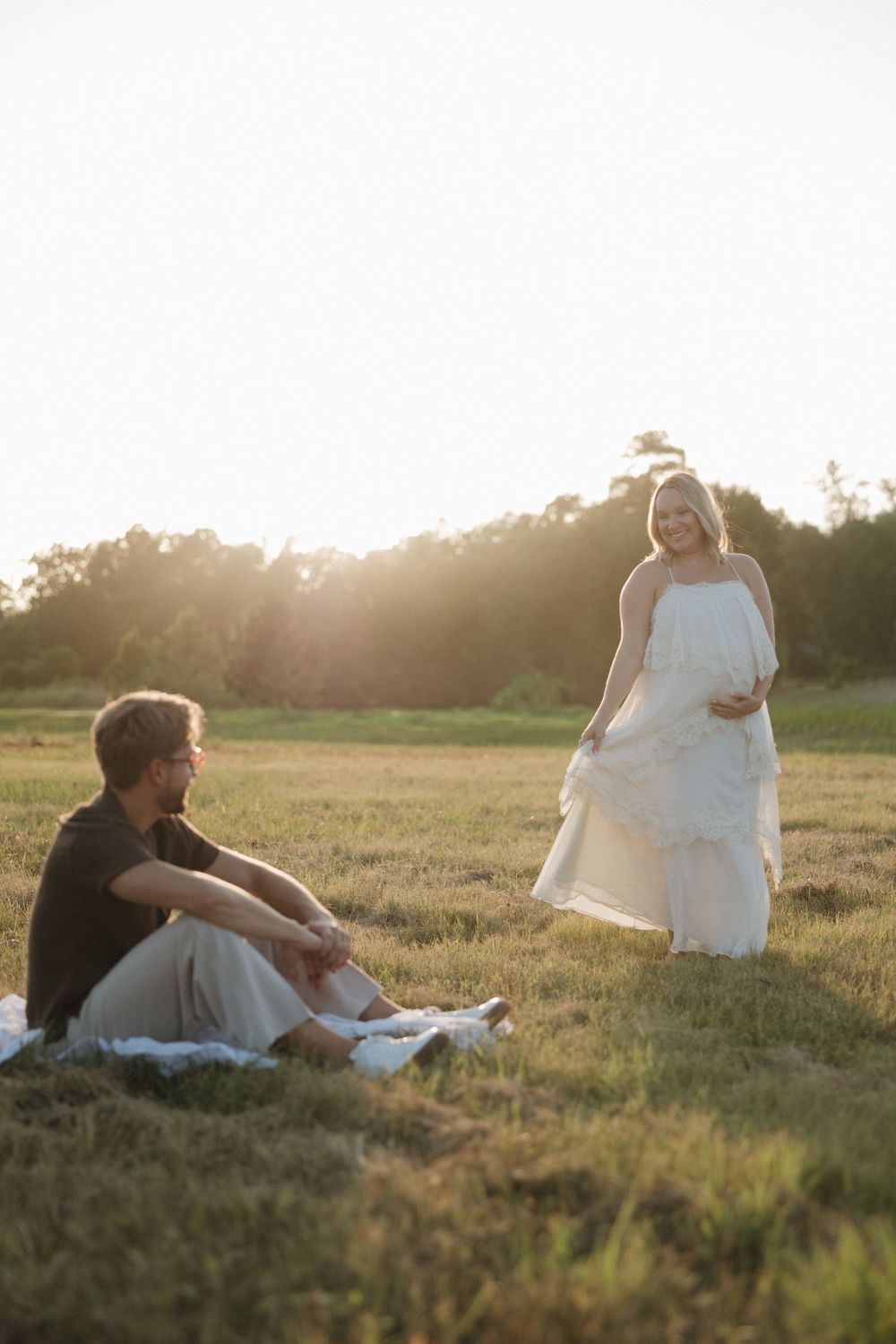 Pregnant woman in white dress stands in sunlit field while man sits on blanket during golden hour maternity photo session.