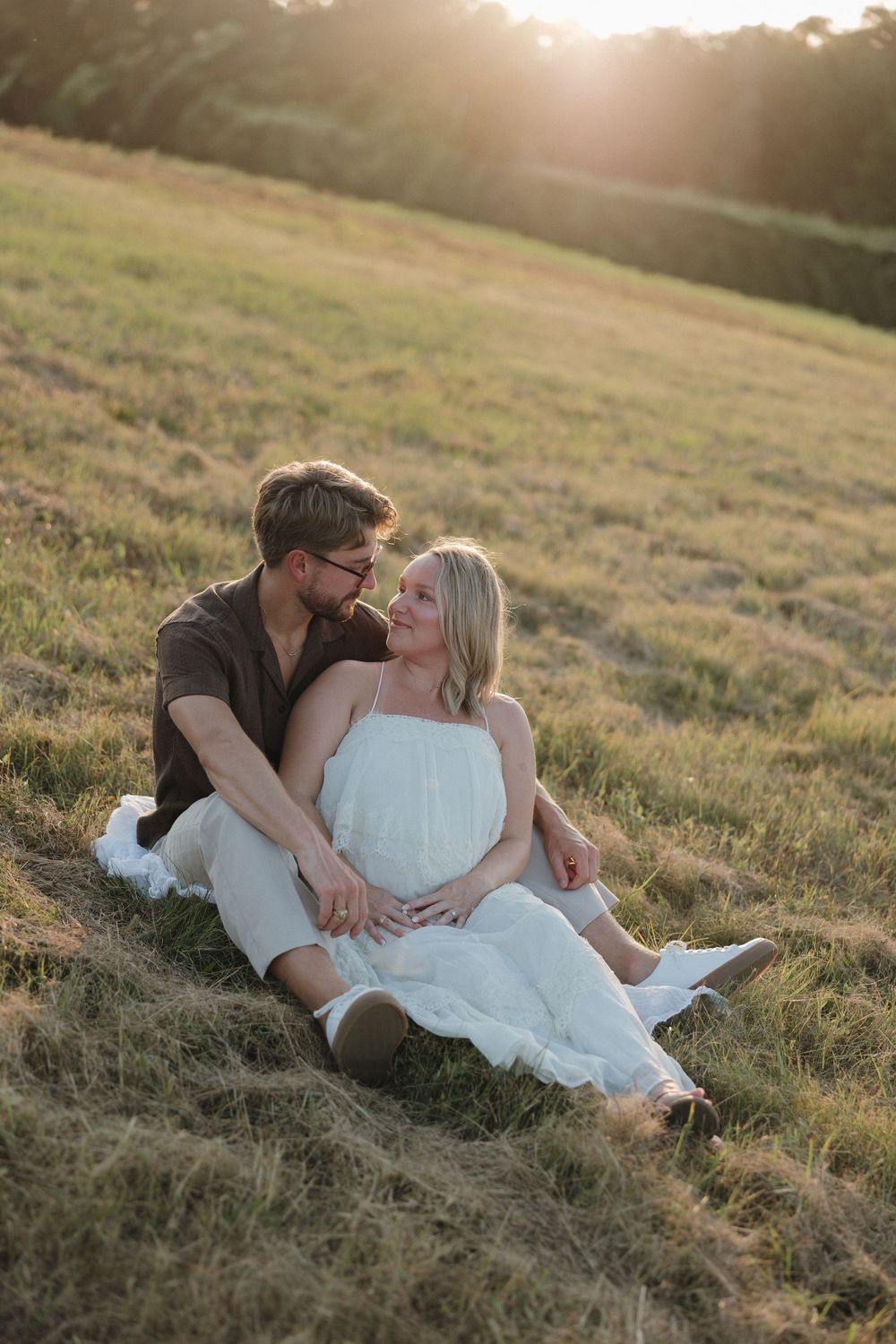 Couple sitting together in a sunlit grassy field at golden hour, woman in white dress leaning against man in brown shirt.
