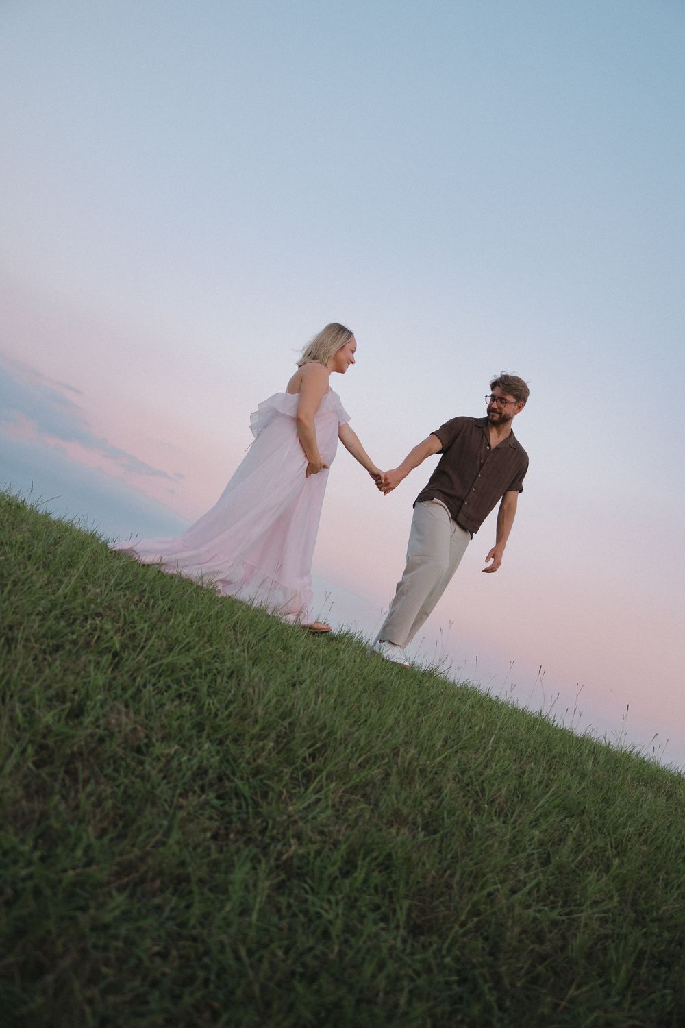 Couple holding hands on a grassy hill at sunset, woman in flowing pink gown, man in casual attire.