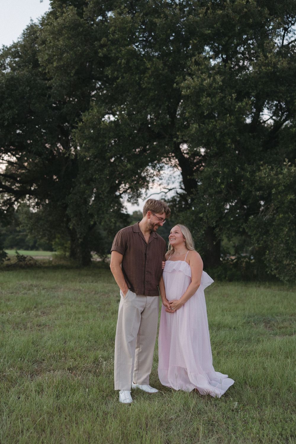 Couple poses in green field, pregnant woman in pink gown with man in brown shirt, large trees backdrop.