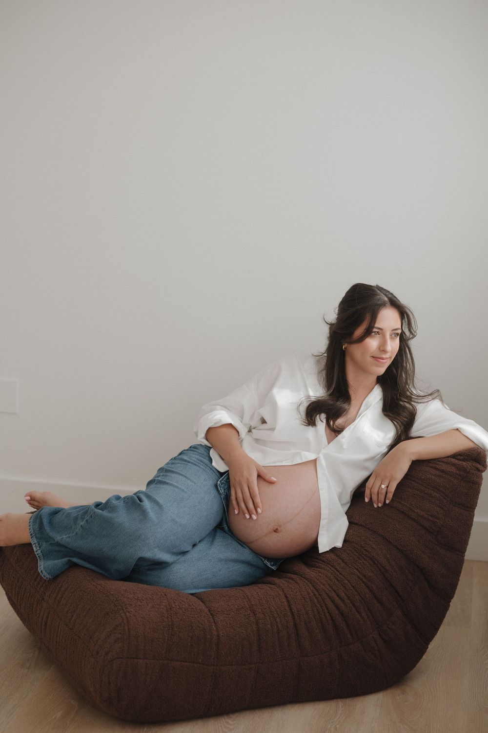 Pregnant woman in white shirt and jeans relaxing on brown bean bag chair, cradling baby bump.