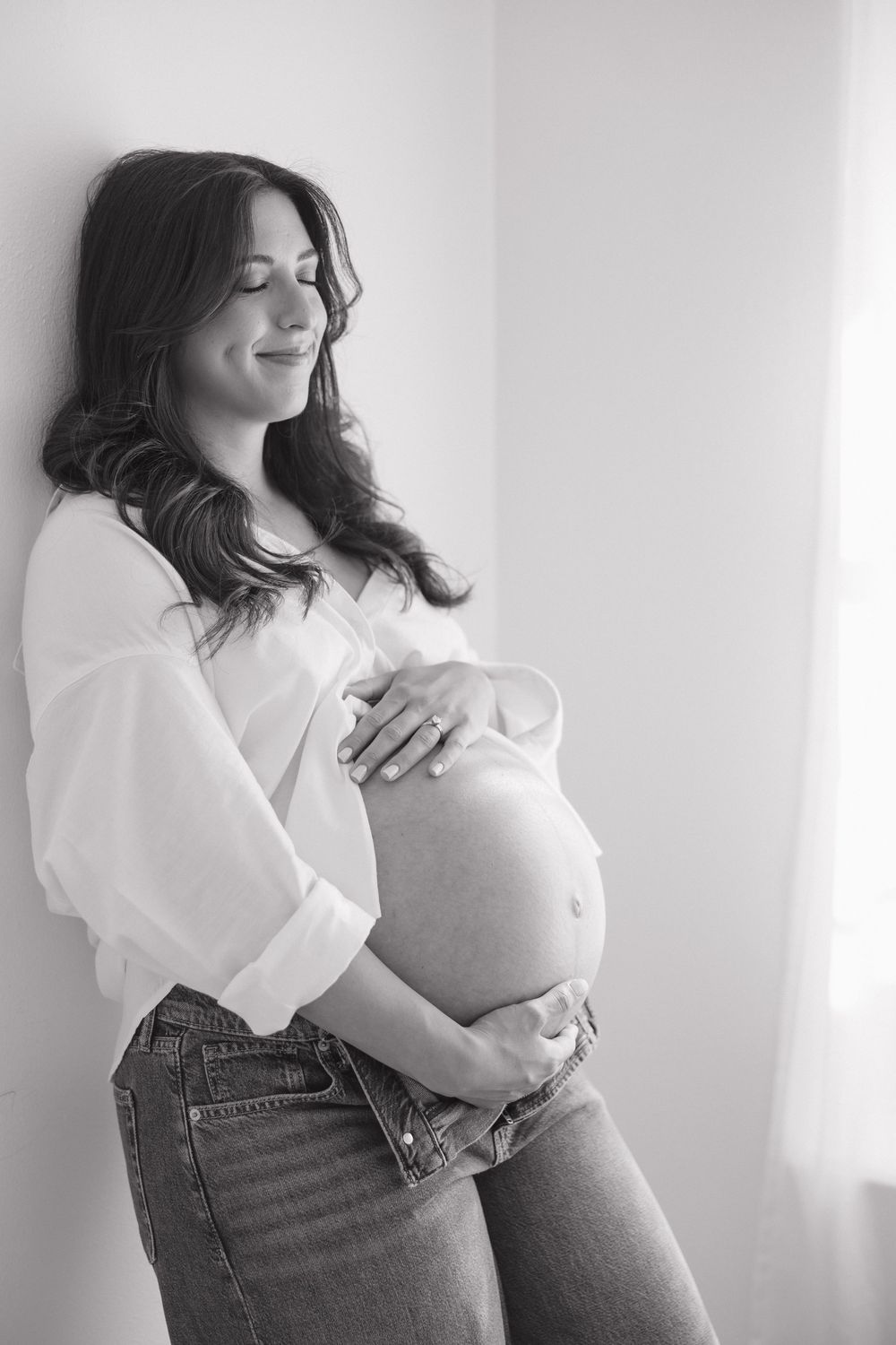 Black and white photo of a smiling pregnant woman in a white shirt and jeans cradling her baby bump against a wall.