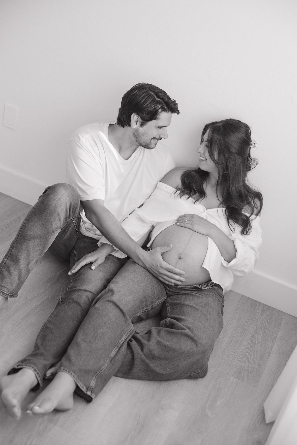 Black and white photo of a couple sitting on floor, man embracing pregnant woman as they smile at each other.